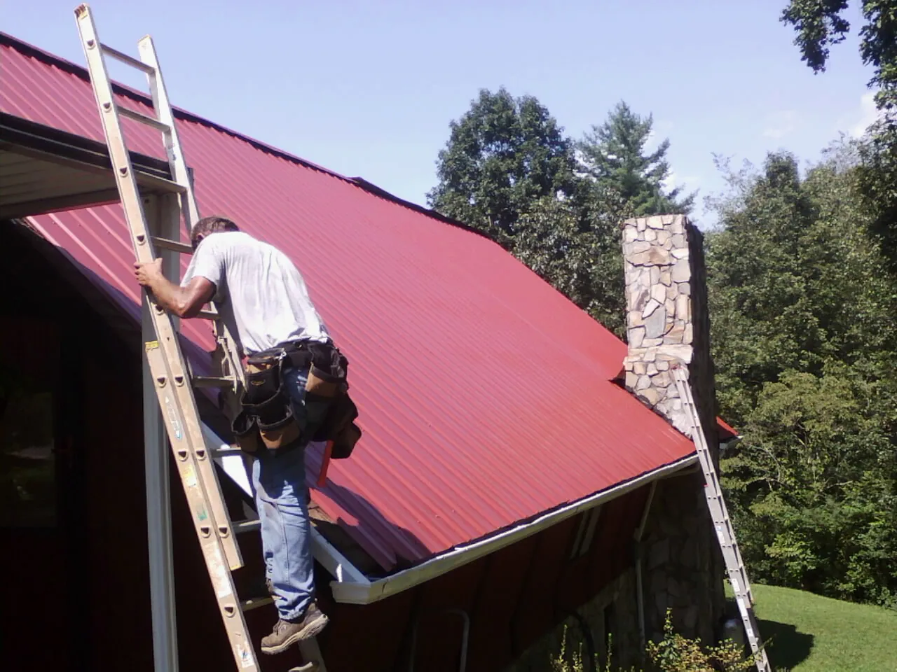 Roofer on a ladder beside a red metal roof and stone chimney — Soffit & Fascia Repair by Roofing Brothers in Burkburnett